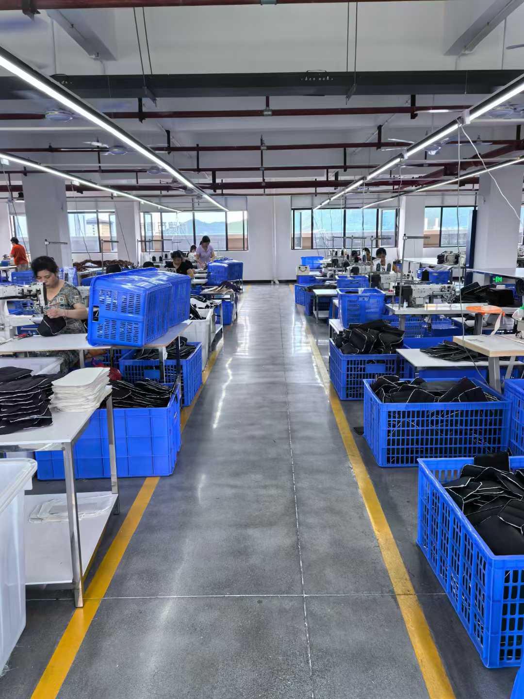 Workers stitching black bag components beside blue baskets containing prepared pieces.