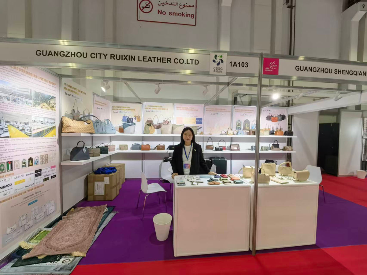 A person presenting handbag and accessory samples behind an exhibition counter.
